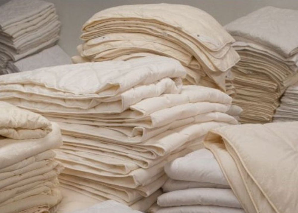 Stacks of folded cream-colored bedding, including duvet covers and quilted blankets, arranged on a bed in a storage or retail setting.