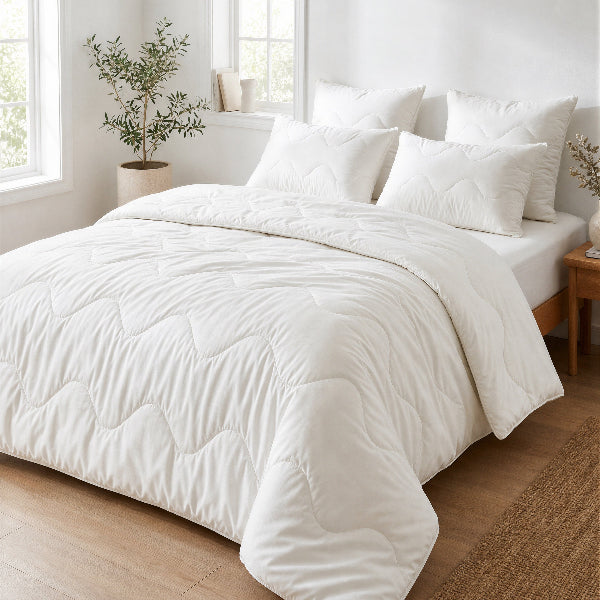 A white quilted comforter with matching pillows and a white sheet on a bed in a bright, modern bedroom with light wood floors and a plant.